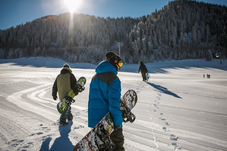 Group of snowboarders walking up a snowy ski slope during a sunny winter day in the mountains.