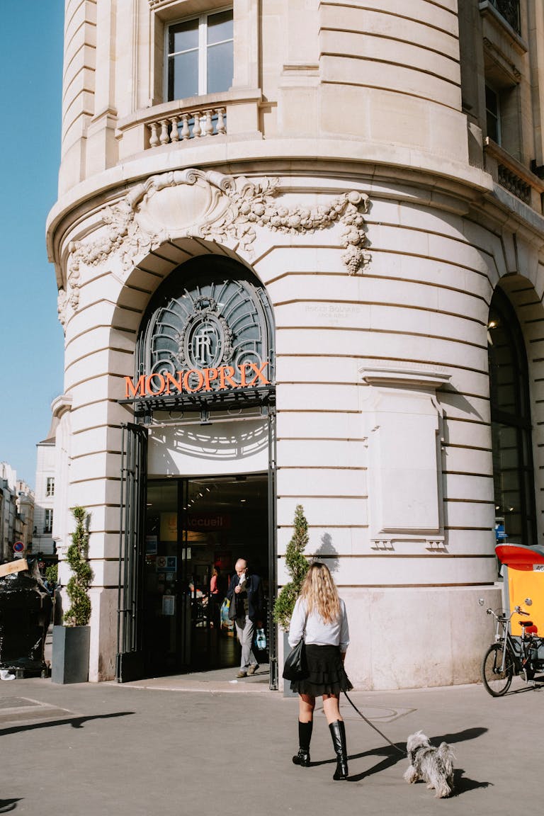 A woman walks her dog outside a historical Monoprix building with a classic architectural design.